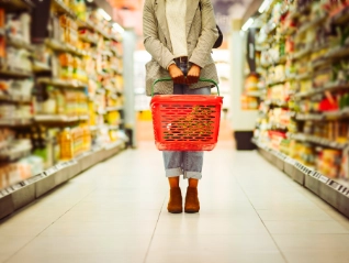 A shopper holding a red plastic shopping basket stands in the brightly lit aisle of a supermarket between two long,...
