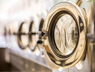  Close-up of a row of commercial washing machine doors in a laundromat, with the first chrome door open and the others...