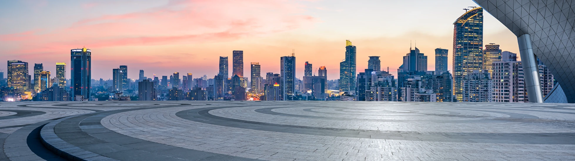 Modern city skyline with high-rises at dusk, viewed from an empty, patterned plaza. A large, curved roof element frames the upper right.