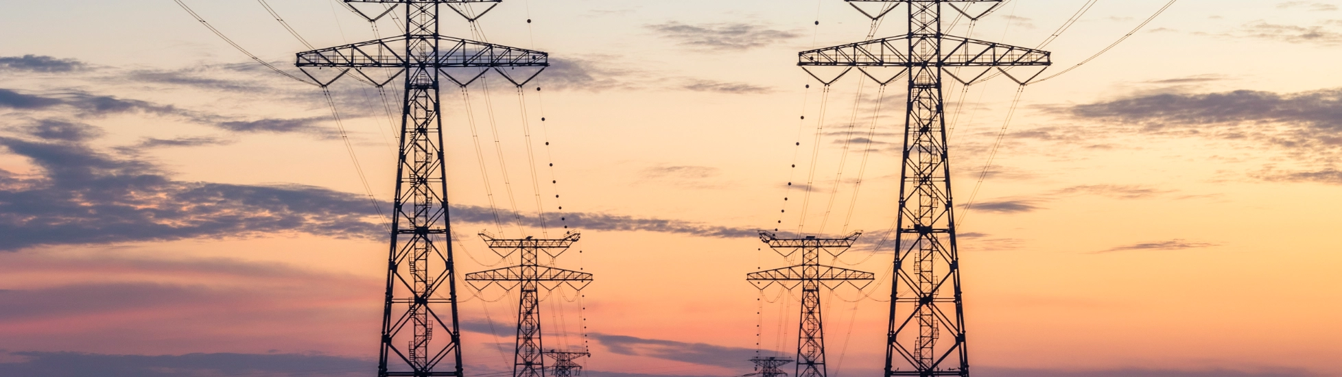 Large metal pylons carry power lines over water, silhouetted against a dramatic sunset of blue to orange.
