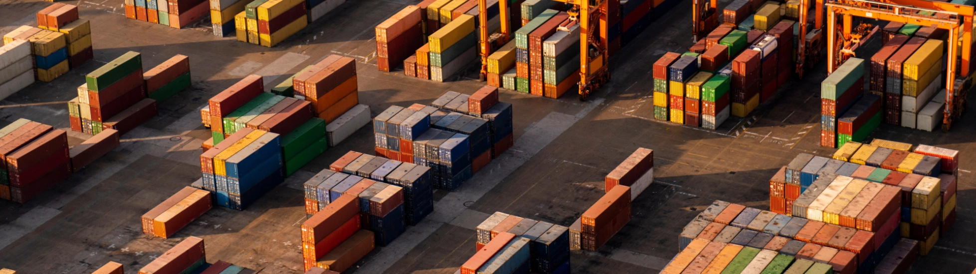 Aerial view: Hundreds of brightly colored shipping containers are stacked high at a busy port/terminal, with large yellow gantry cranes overhead.